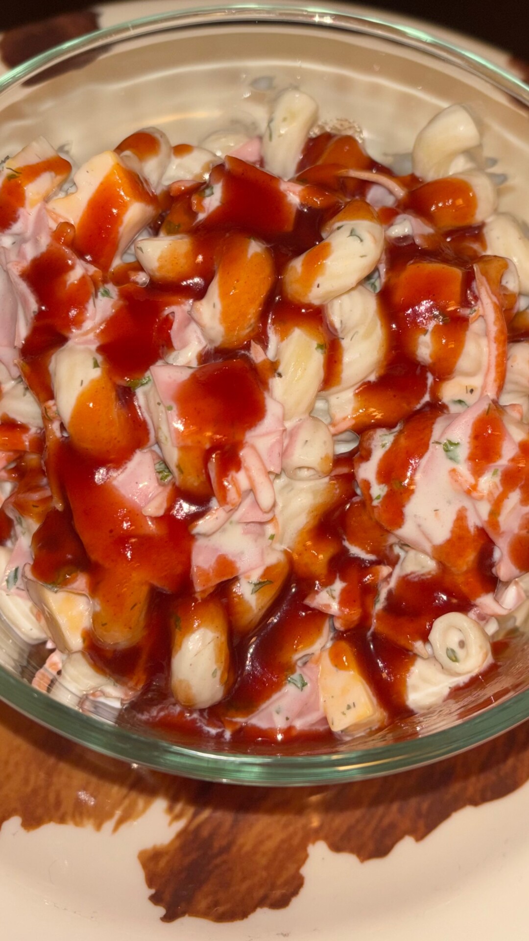 A glass bowl filled with pasta salad mixed with diced ham, carrots, and herbs, topped with a generous drizzle of dark red sauce.