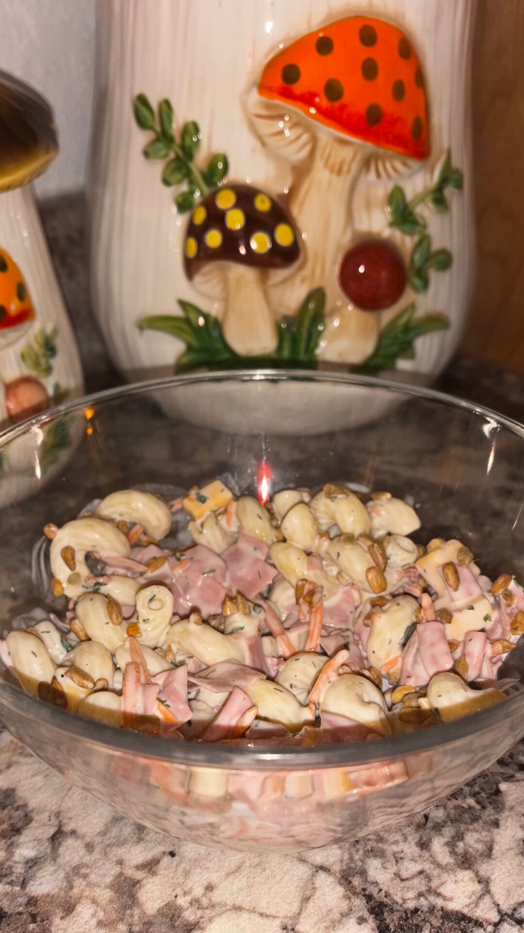A glass bowl of pasta salad with ham and vegetables sits on a counter, with ceramic canisters featuring colorful mushrooms in the background.