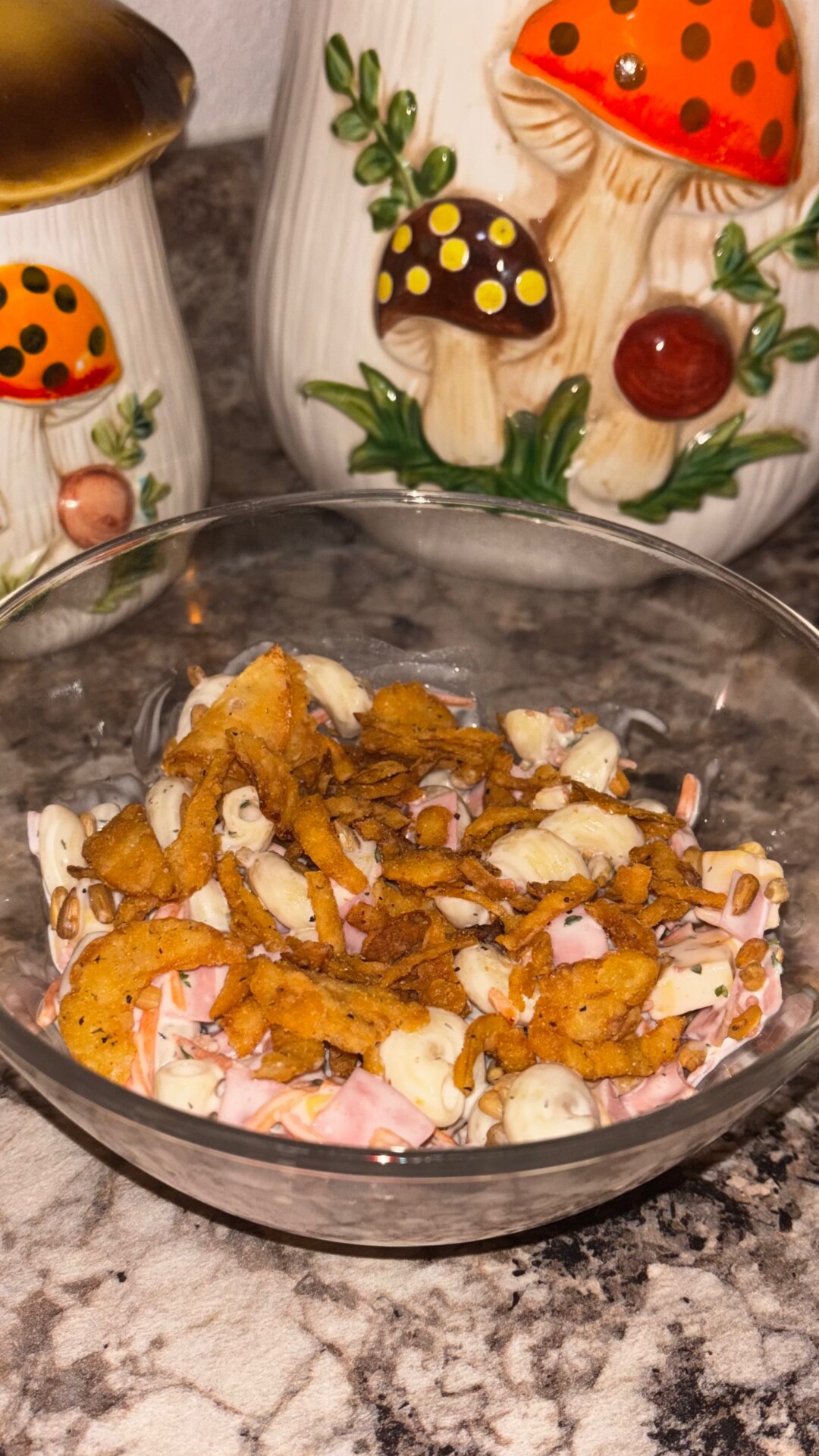 A glass bowl with a salad topped with crushed chips sits on a marble counter. In the background, there are decorative ceramic containers with painted mushrooms.