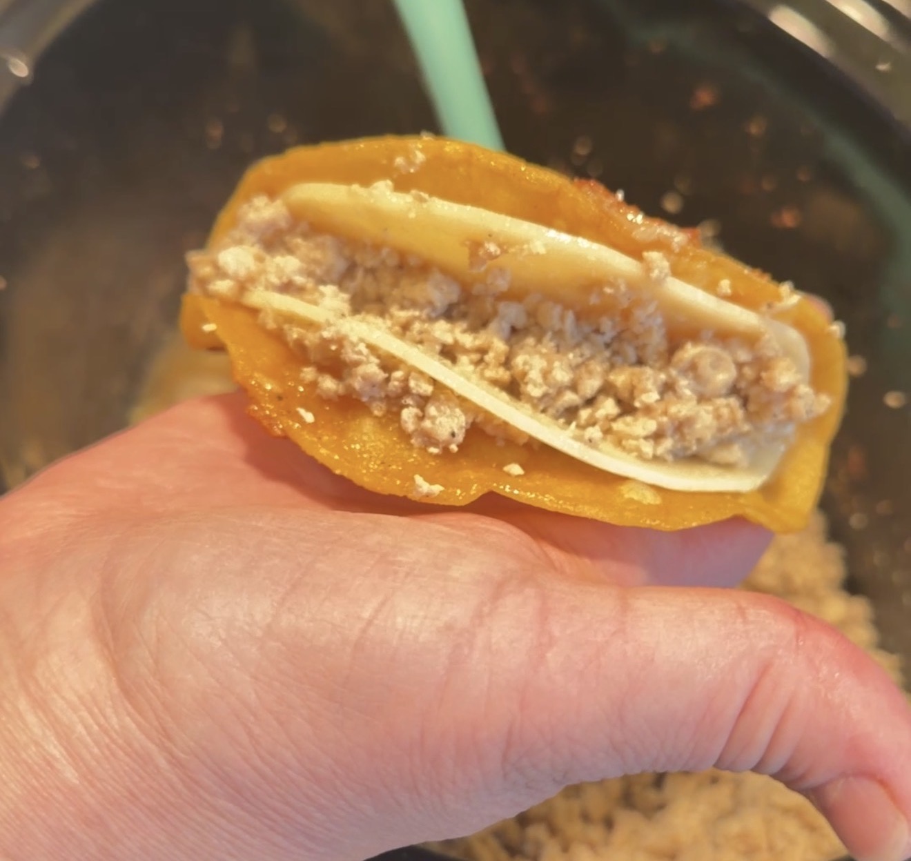 A hand holds a yellow taco shell filled with crumbled ground meat and a layer of white cheese, with a blurry background showing more filling in a bowl.
