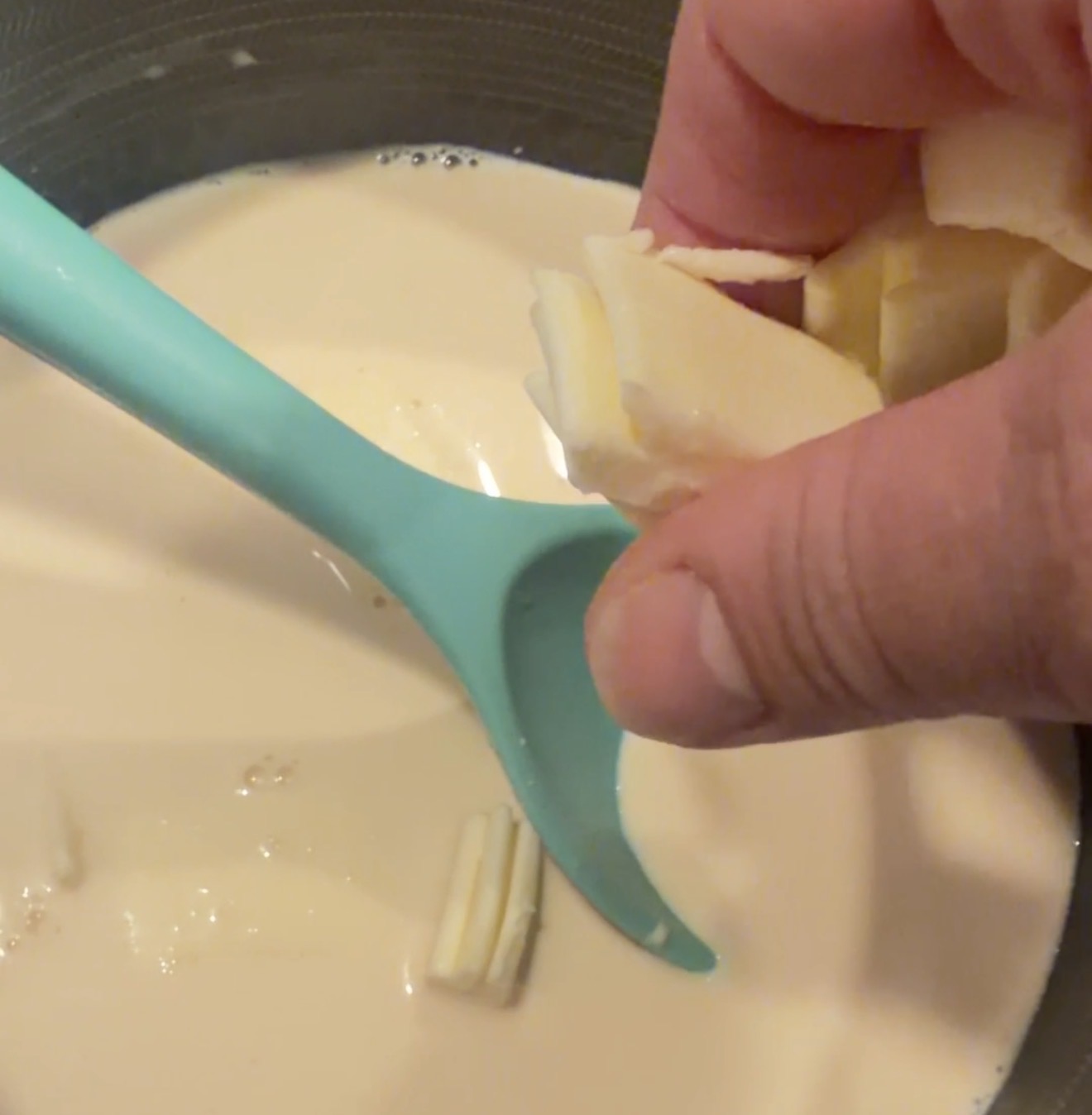 A hand drops slices of butter into a pot of cream while stirring with a blue spoon.