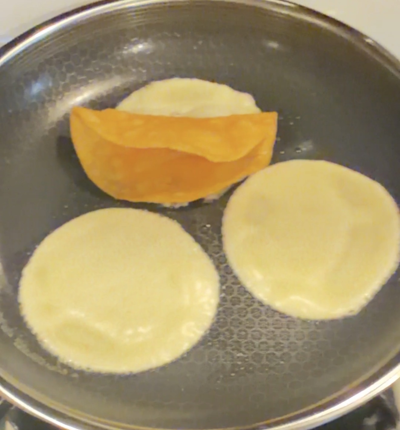 Three corn tortillas cooking in a pan: one is folded in half and crisp, while the other two are still flat and soft, starting to cook.