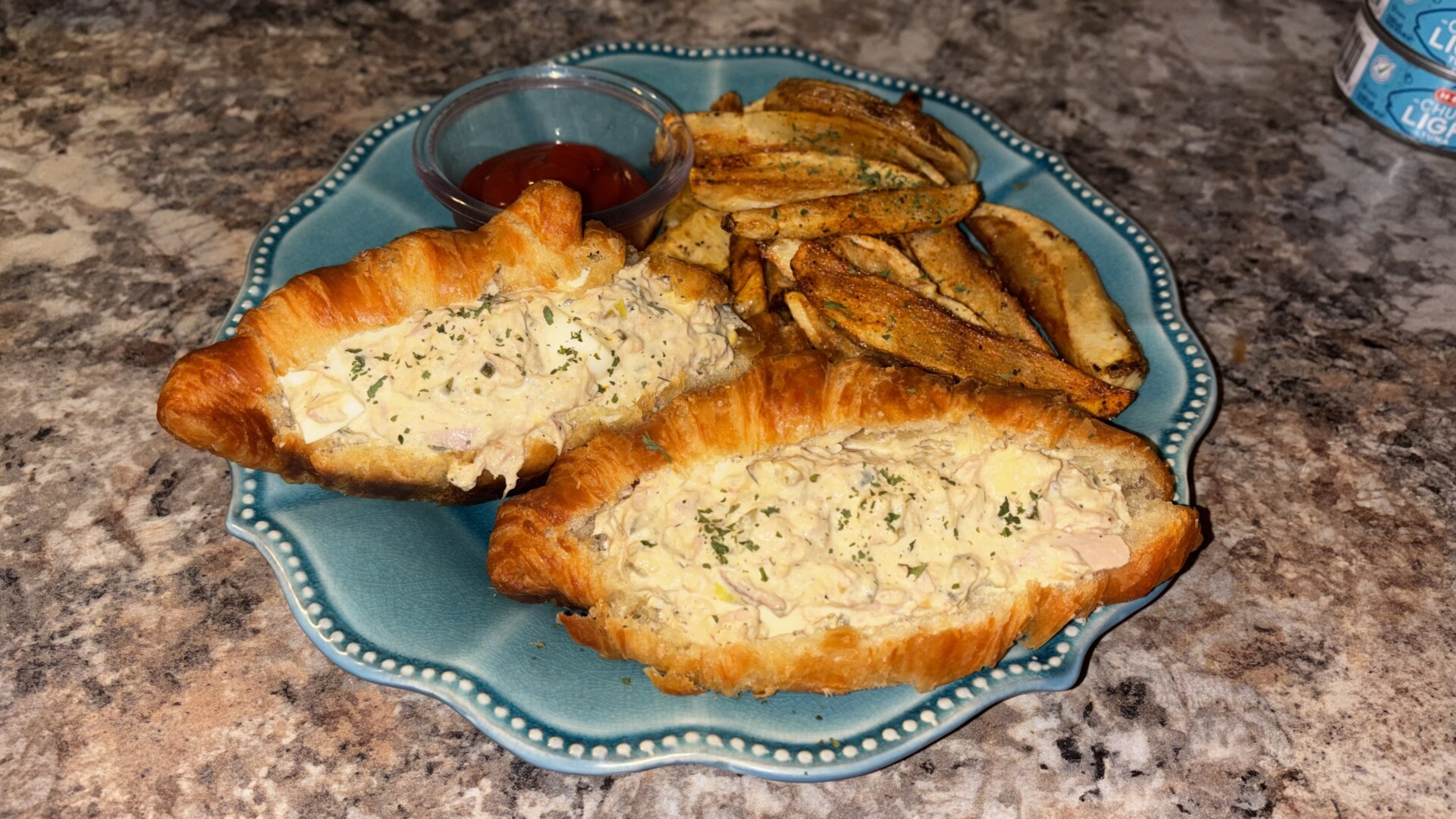 A blue plate with a croissant stuffed with creamy tuna salad, topped with herbs, cut in half, served with seasoned potato wedges and a small container of ketchup on a marble countertop.