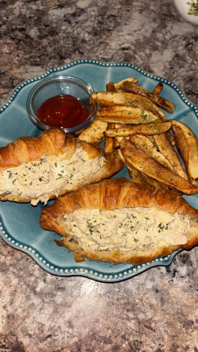 A blue plate with two croissant sandwiches filled with creamy chicken salad, seasoned potato wedges, and a small dish of ketchup on a marbled countertop.