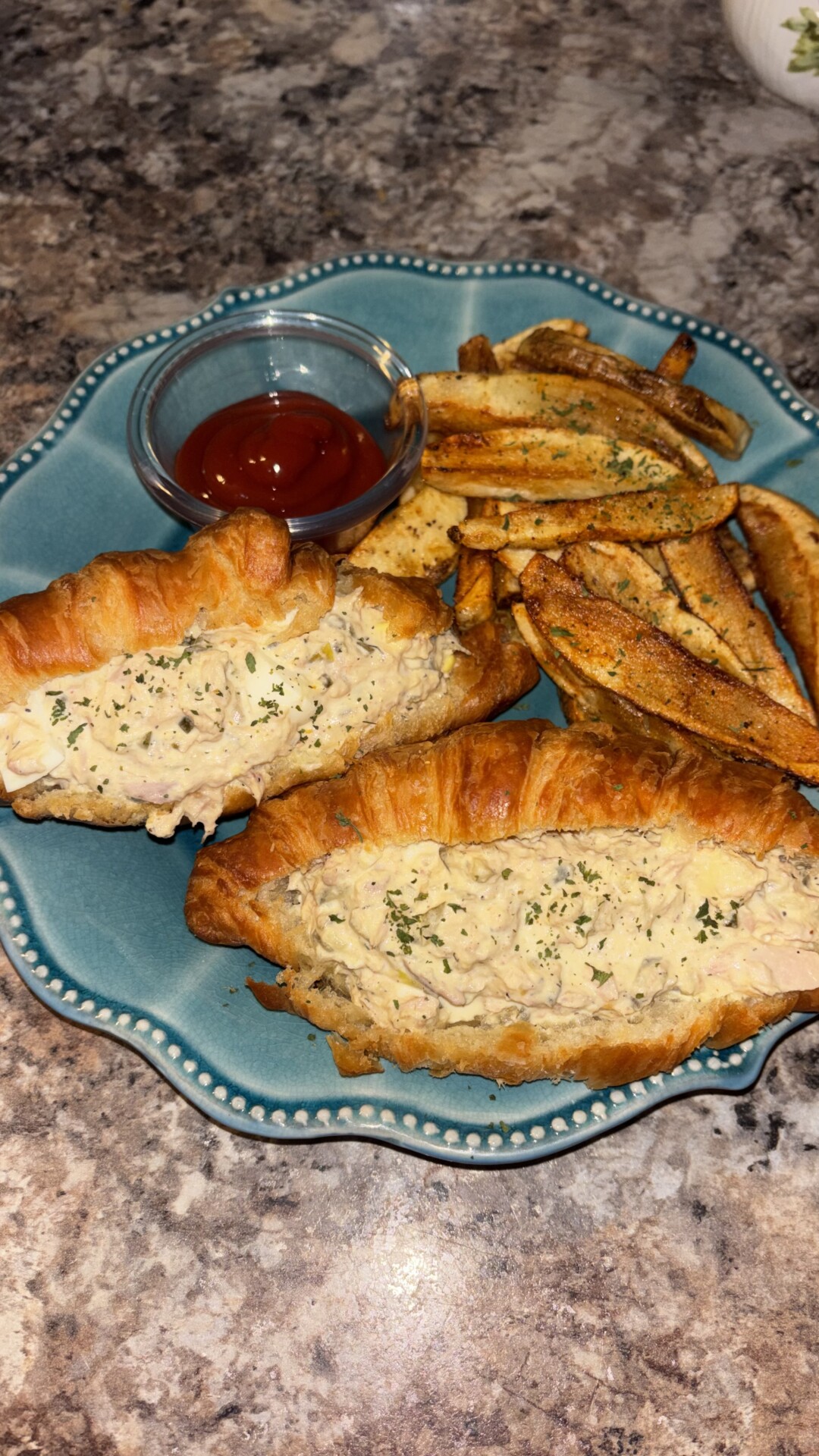 A blue plate with two croissant sandwiches filled with a creamy mixture, seasoned potato wedges, and a small glass dish of ketchup, all on a granite countertop.
