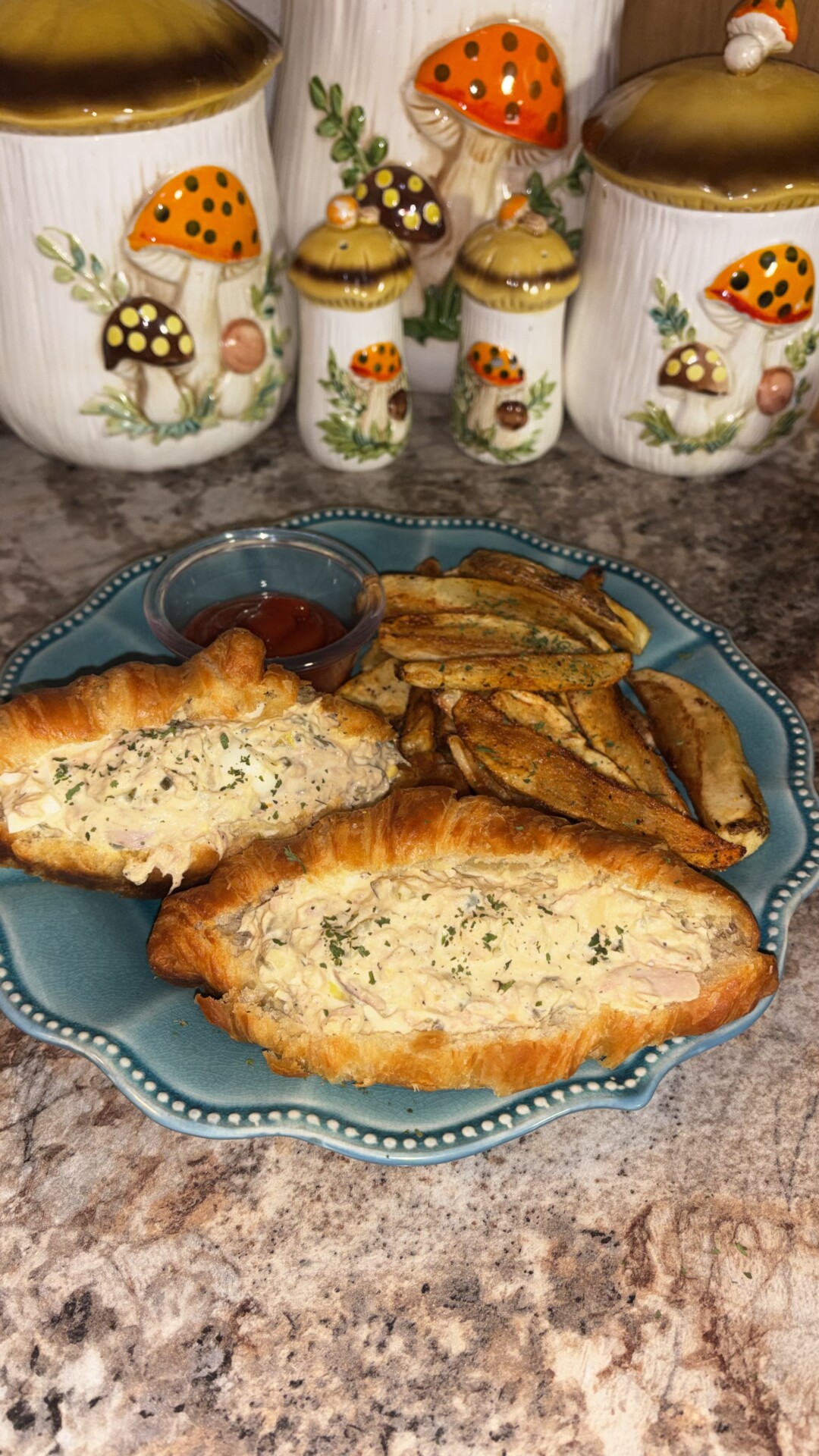 A teal plate with two stuffed croissants, seasoned fries, and a cup of ketchup. In the background, there are ceramic containers decorated with colorful mushrooms on a kitchen counter.
