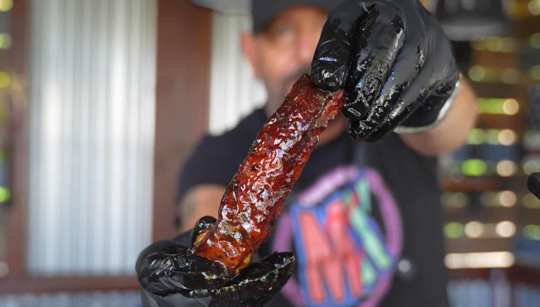 A person wearing black gloves holds up a glazed, grilled meat rib toward the camera. The background is blurred, and the person’s shirt has a colorful circular design.