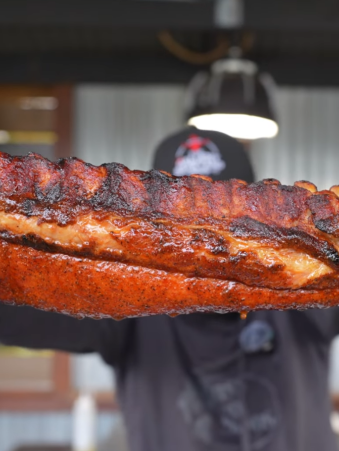 A person holds up a slab of grilled, seasoned ribs in front of them, with the focus on the meat and a blurred background of an outdoor setting.