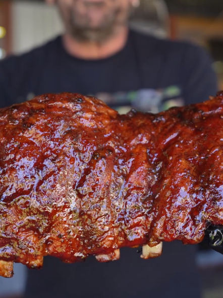 A person holds a rack of glazed, barbecued ribs close to the camera, showing the shiny, caramelized sauce on the meat. The person’s face is blurred in the background.