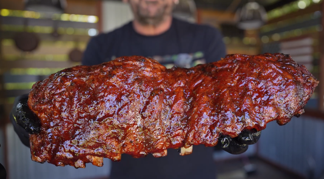A person wearing black gloves holds out a large rack of glazed barbecue ribs, with a shiny, saucy coating. The background is blurred, and the ribs are the main focus of the image.