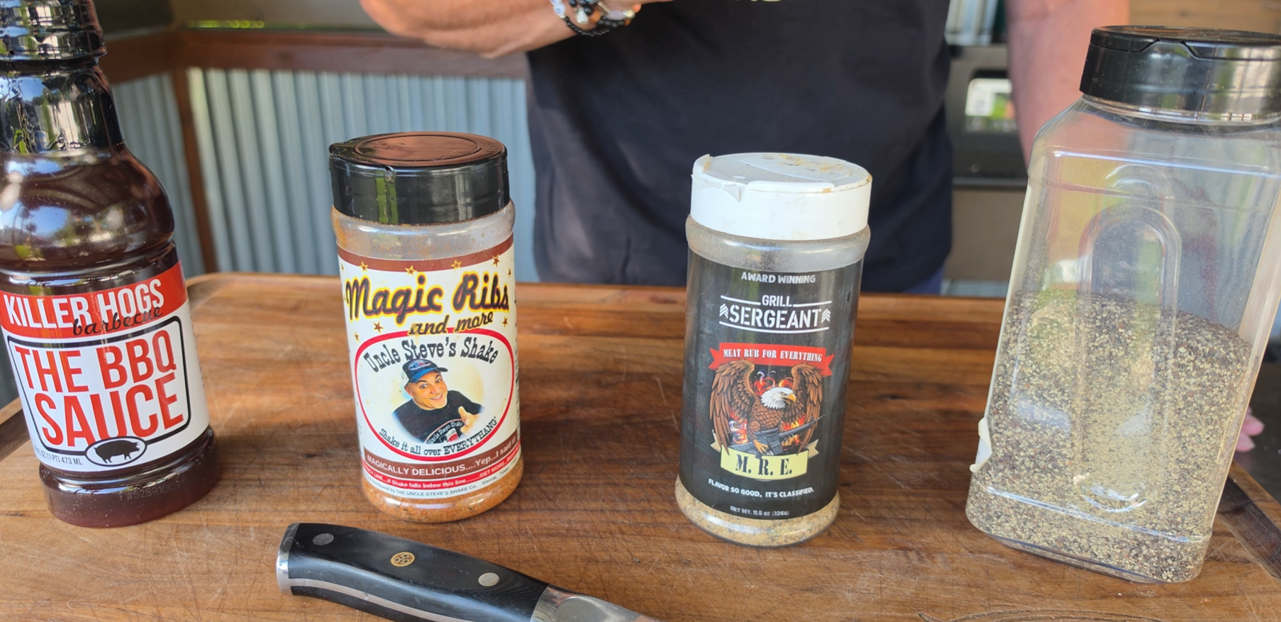 Four seasoning containers and a knife on a wooden table: Killer Hogs BBQ Sauce, Magic Dust, Drill Sergeant seasoning, and a plastic container of black pepper. A person stands in the background.