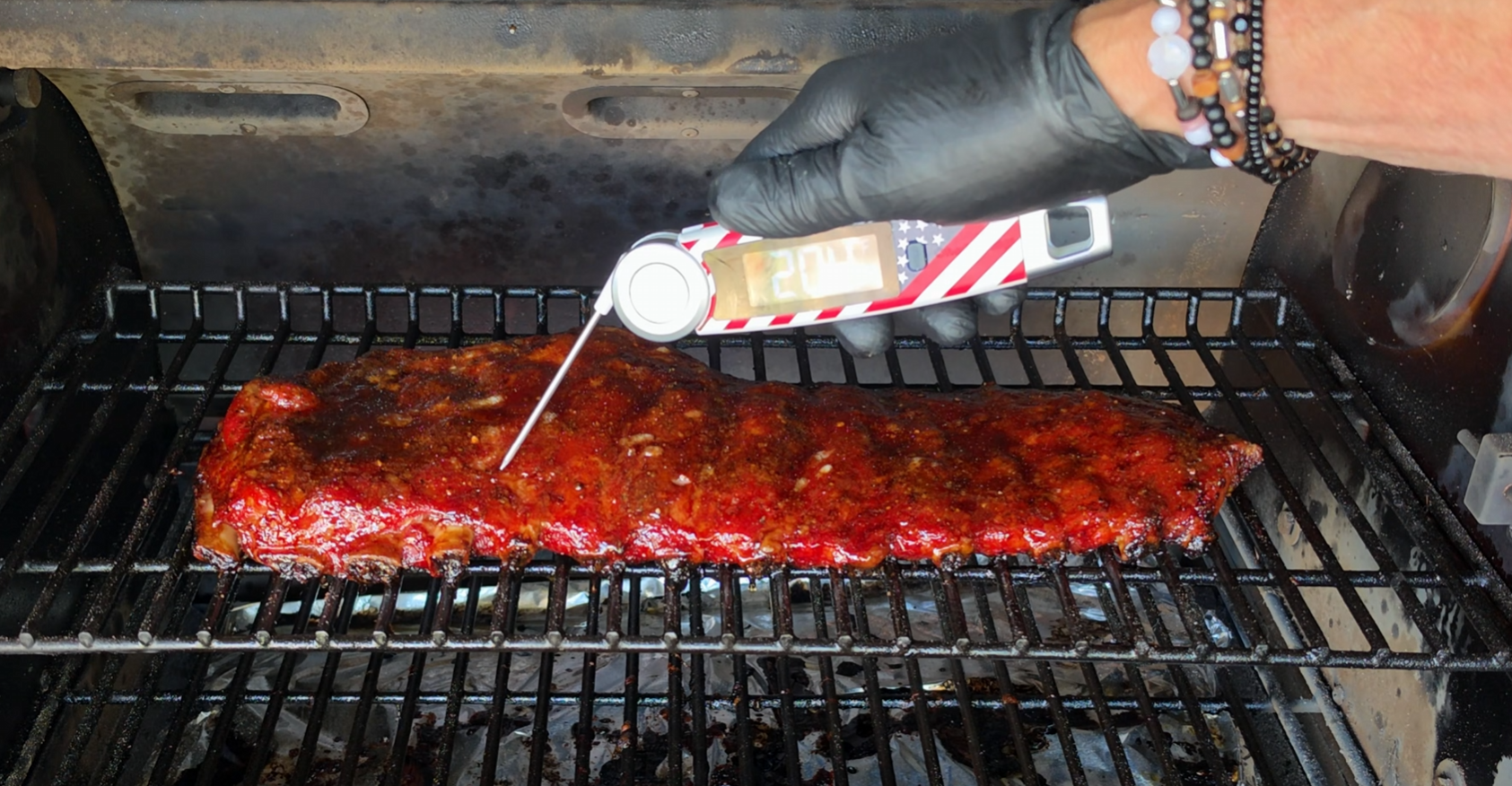 A hand wearing a black glove uses a meat thermometer to check ribs cooking on a grill. The thermometer reads 200°F. The ribs have a glossy, seasoned exterior.
