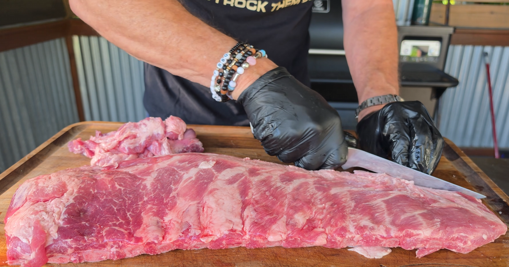 Person wearing black gloves trims a large slab of raw pork ribs on a wooden cutting board with a knife. A grill and kitchen items are visible in the background.
