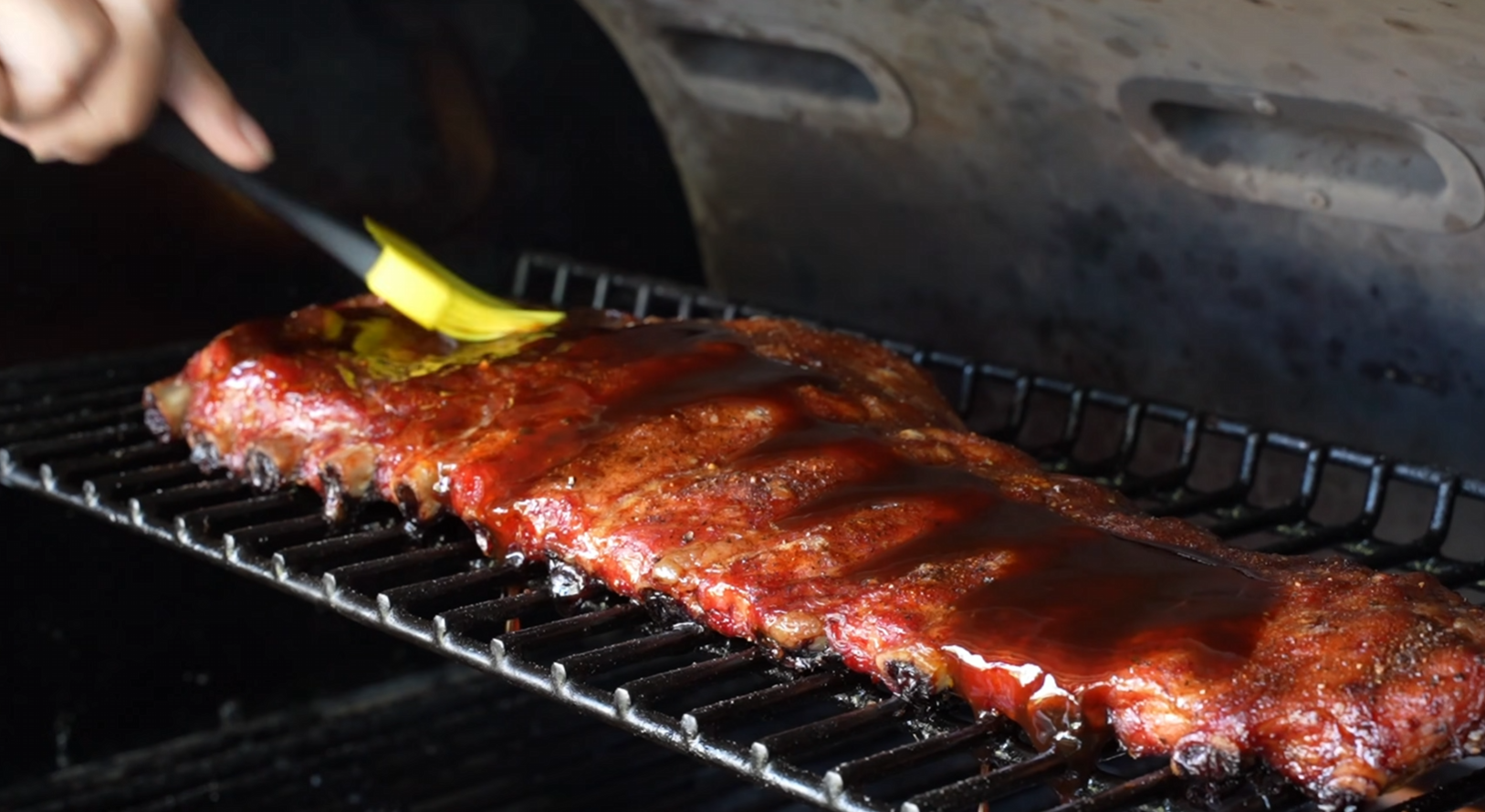 A rack of ribs on a grill is being brushed with sauce using a yellow brush, with smoke or heat visible from the grill.