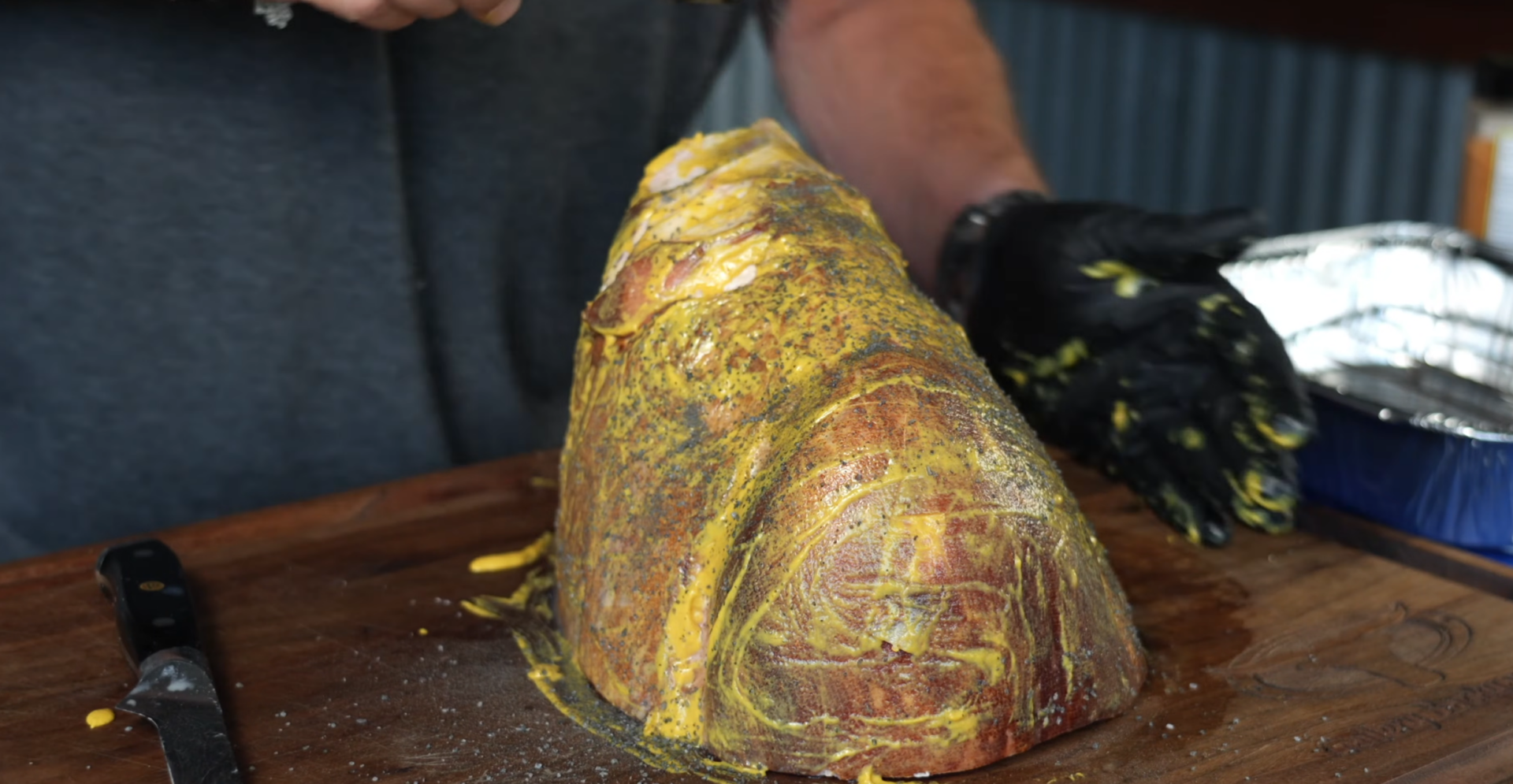 A person in gloves spreads mustard and seasoning on a large cut of meat on a wooden surface. A knife and a foil tray are nearby.