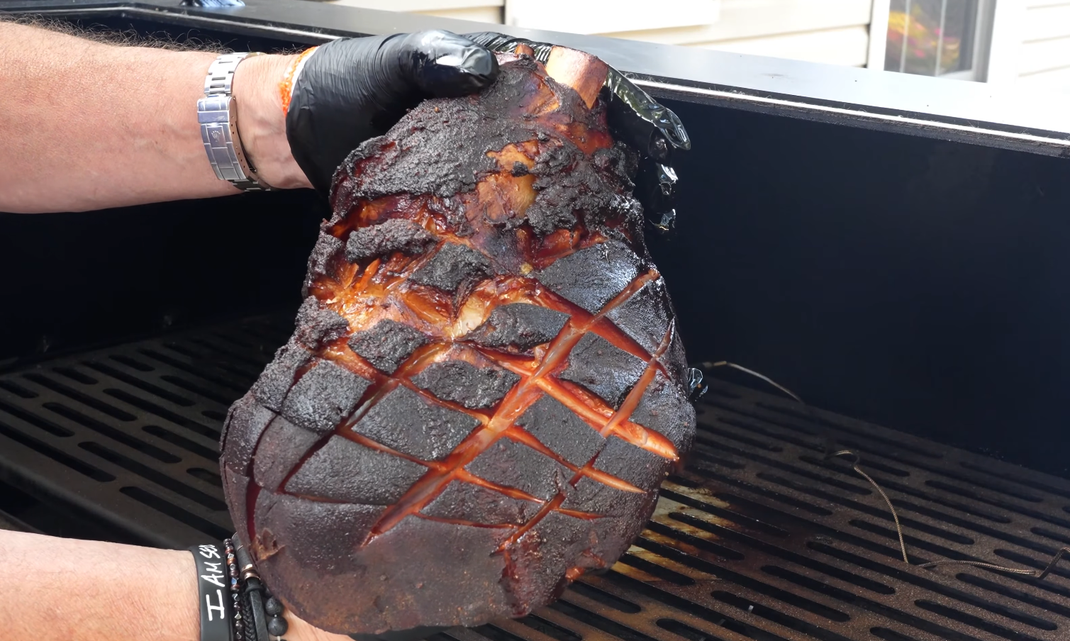 A person wearing black gloves holds a large, smoked ham with a diamond pattern cut into the surface over a grill. The ham is dark and caramelized from cooking.
