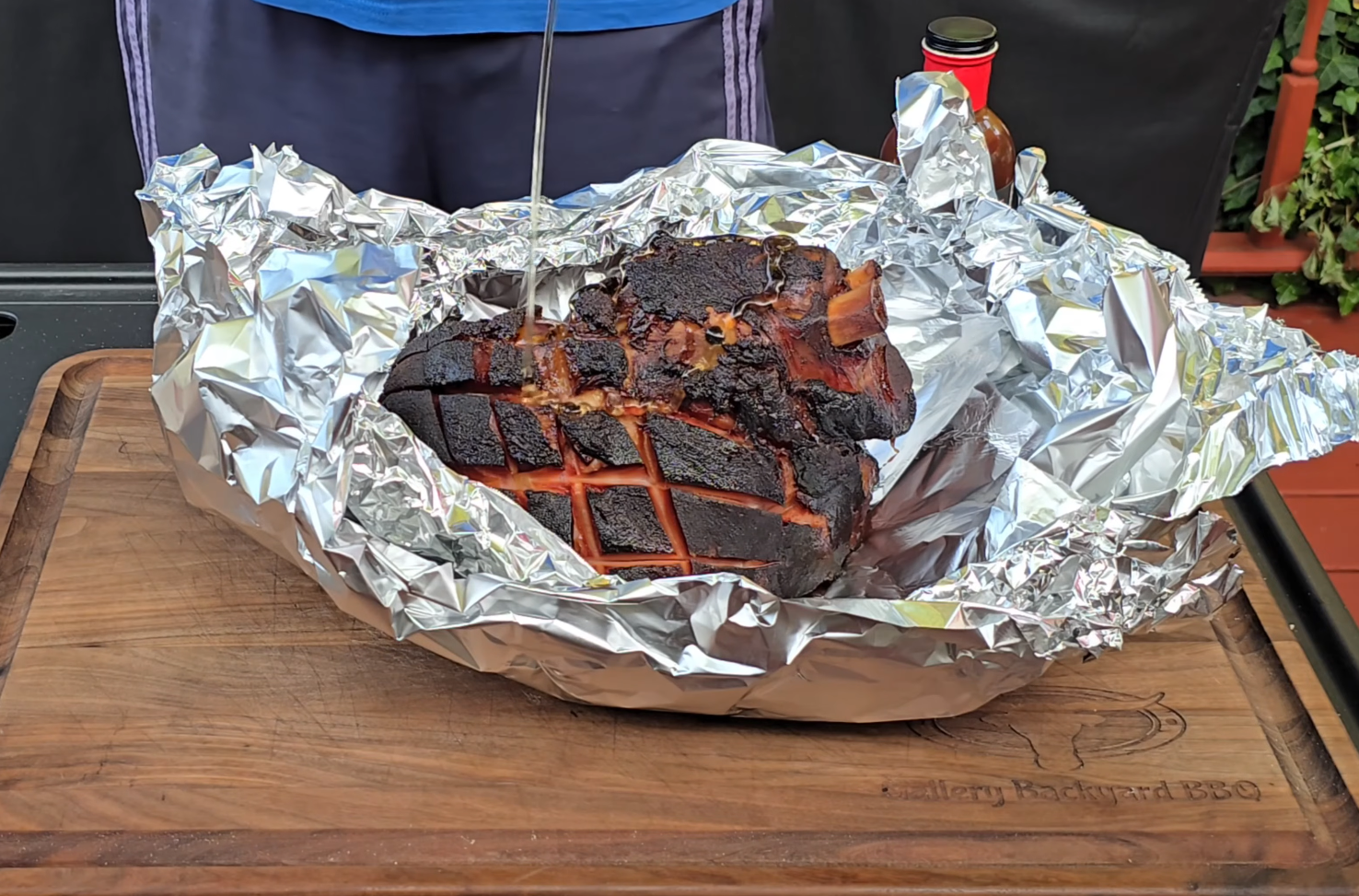 A smoked ham with a crispy, dark crust sits on a sheet of aluminum foil atop a wooden cutting board, with a person standing behind it.