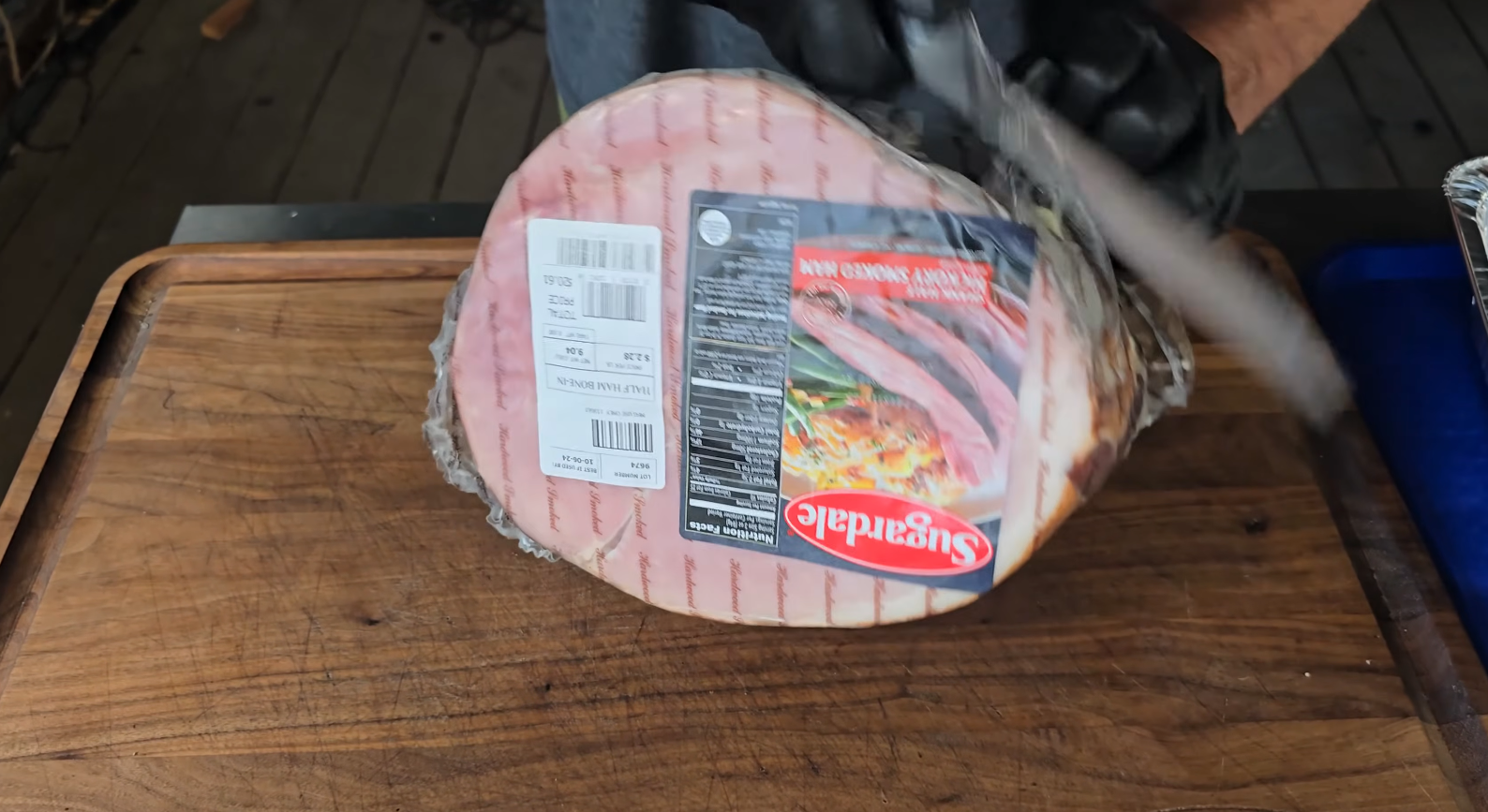 A person wearing black gloves slices a large Sugardale spiral sliced ham on a wooden cutting board. The ham is still partially wrapped in plastic with a visible label.