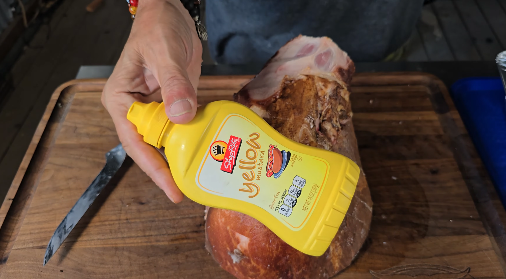 A hand holds a yellow squeeze bottle of Siefa yellow mustard over a cooked ham on a wooden cutting board; a knife is nearby.