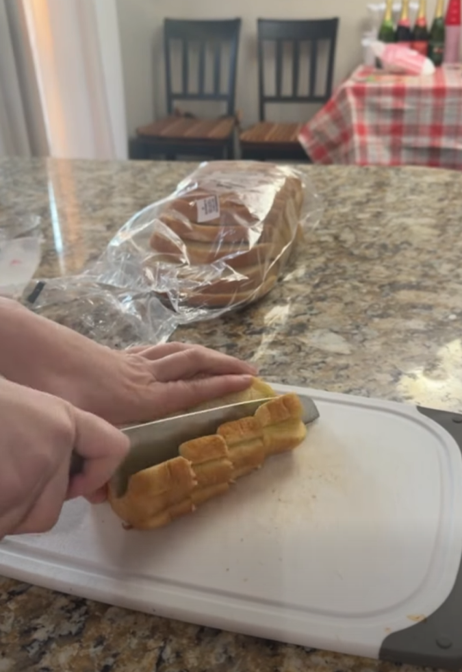 A person slices a loaf of bread on a white cutting board, with a bag of sliced bread and a table with a red checkered tablecloth visible in the background.