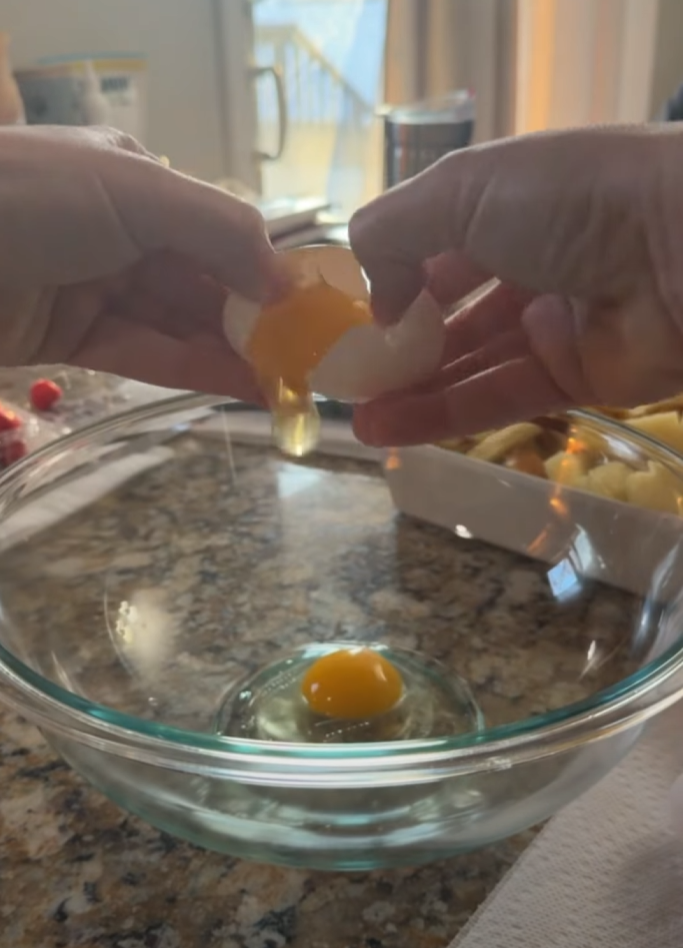 Two hands crack an egg above a glass bowl on a kitchen counter, with the yolk and egg white falling in. There are various kitchen items and food visible in the background.