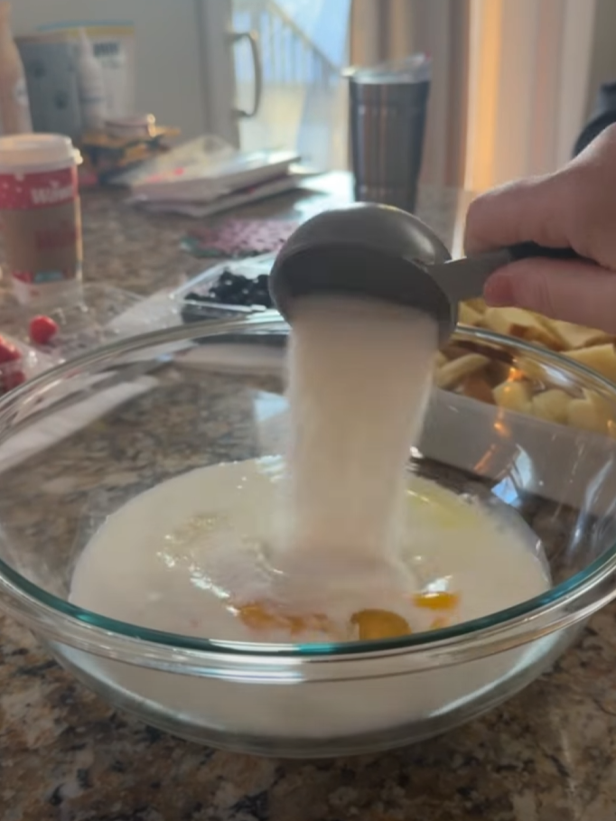 A hand pours a cup of sugar into a glass bowl containing eggs and milk on a kitchen counter, with fruit and various kitchen items in the background.