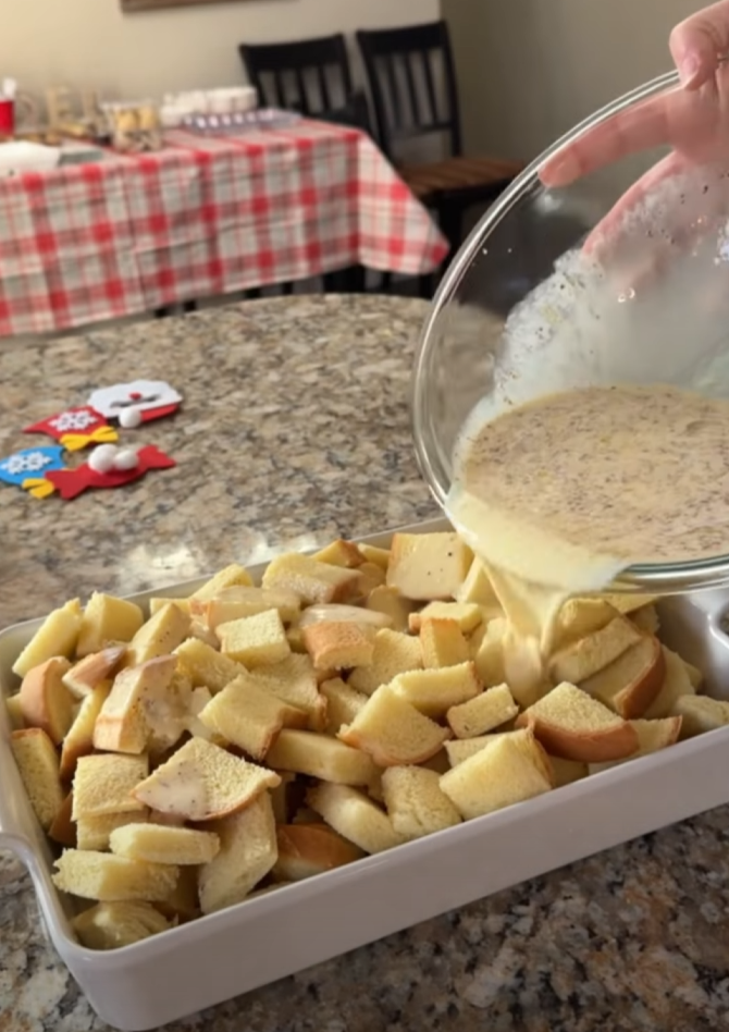 A hand pours a mixture over cubed bread in a baking dish on a kitchen counter. In the background, a table with a red checkered tablecloth and holiday decorations is visible.