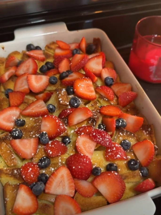 A baked casserole dish topped with sliced strawberries and whole blueberries sits next to a red candle on a kitchen counter.