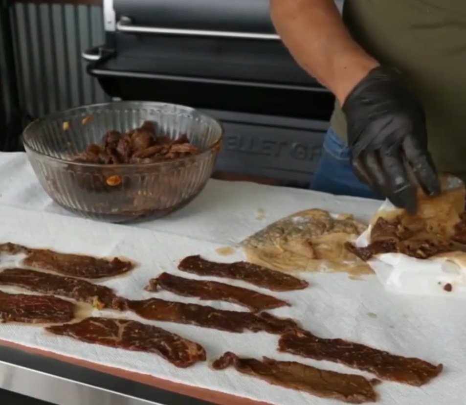 A person wearing a black glove arranges strips of marinated meat on paper towels, with a large bowl of marinated meat and a grill visible in the background.