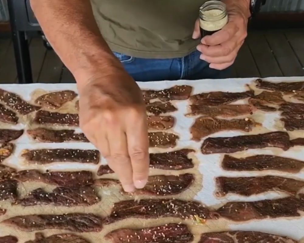 A person sprinkles seasoning onto several rows of raw marinated beef strips laid out on a table, preparing them for cooking or drying.