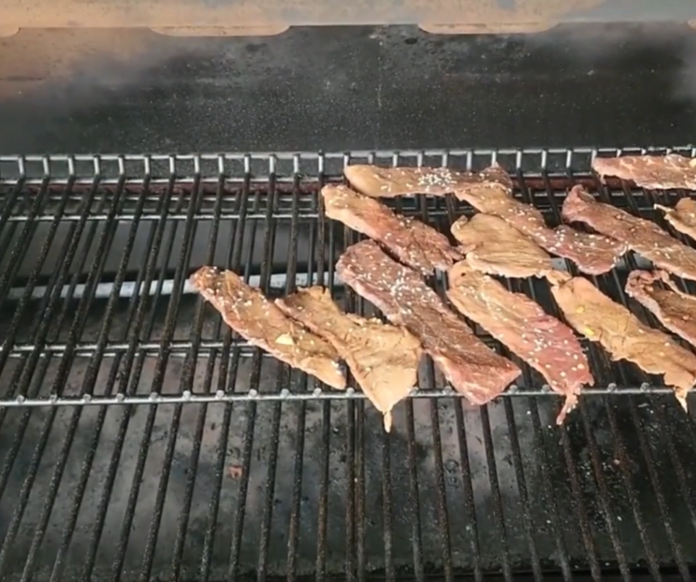 Thin strips of marinated beef are arranged on the top rack of a barbecue grill as they cook, with visible grill marks and smoke rising in the background.