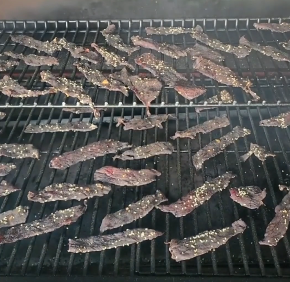 Thin strips of seasoned beef jerky are laid out across two grates inside a grill, being smoked or dried.