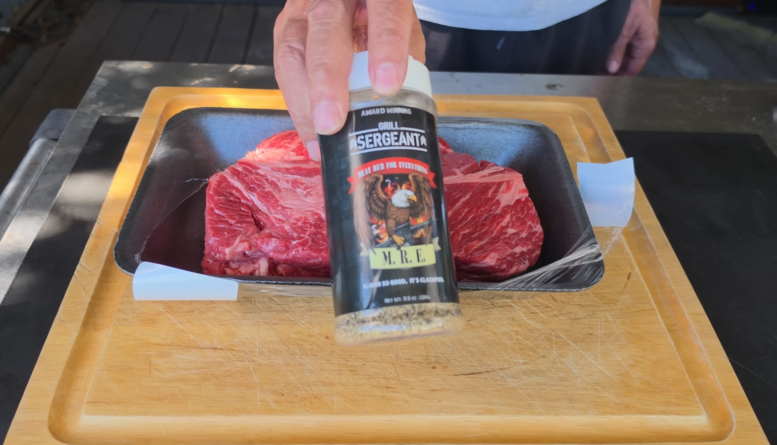 A hand holds a bottle of seasoning labeled Sergeants M.R.E. above a tray of raw beef on a wooden cutting board.