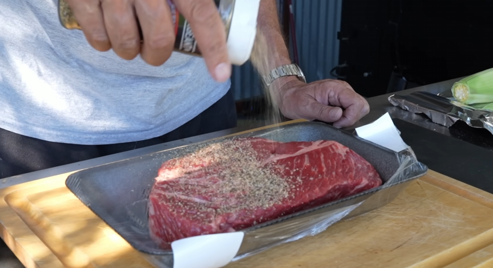 A person sprinkles seasoning onto a raw steak in a tray lined with plastic wrap, preparing it on a wooden cutting board outdoors.