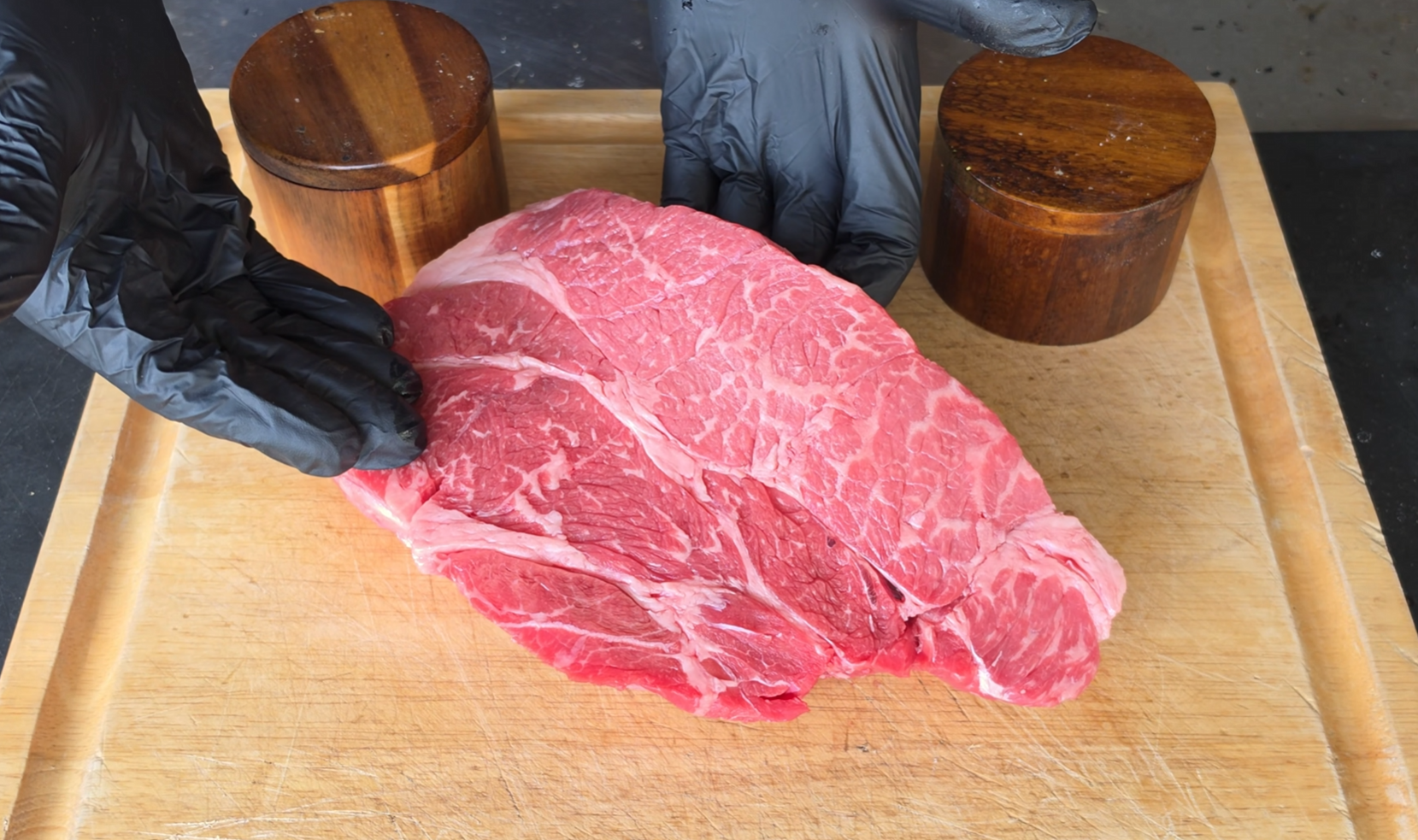 A person wearing black gloves holds a large, raw piece of marbled beef on a wooden cutting board, with two wooden containers nearby.