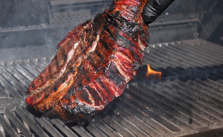 A large, charred rack of beef ribs is being held with tongs over a smoky grill with visible flames and grill marks.