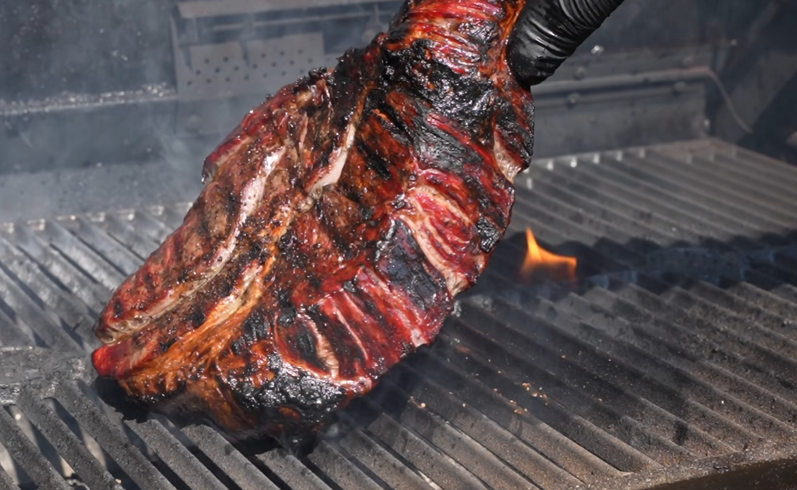A large, charred rack of beef ribs is being held with tongs over a smoky grill with visible flames and grill marks.
