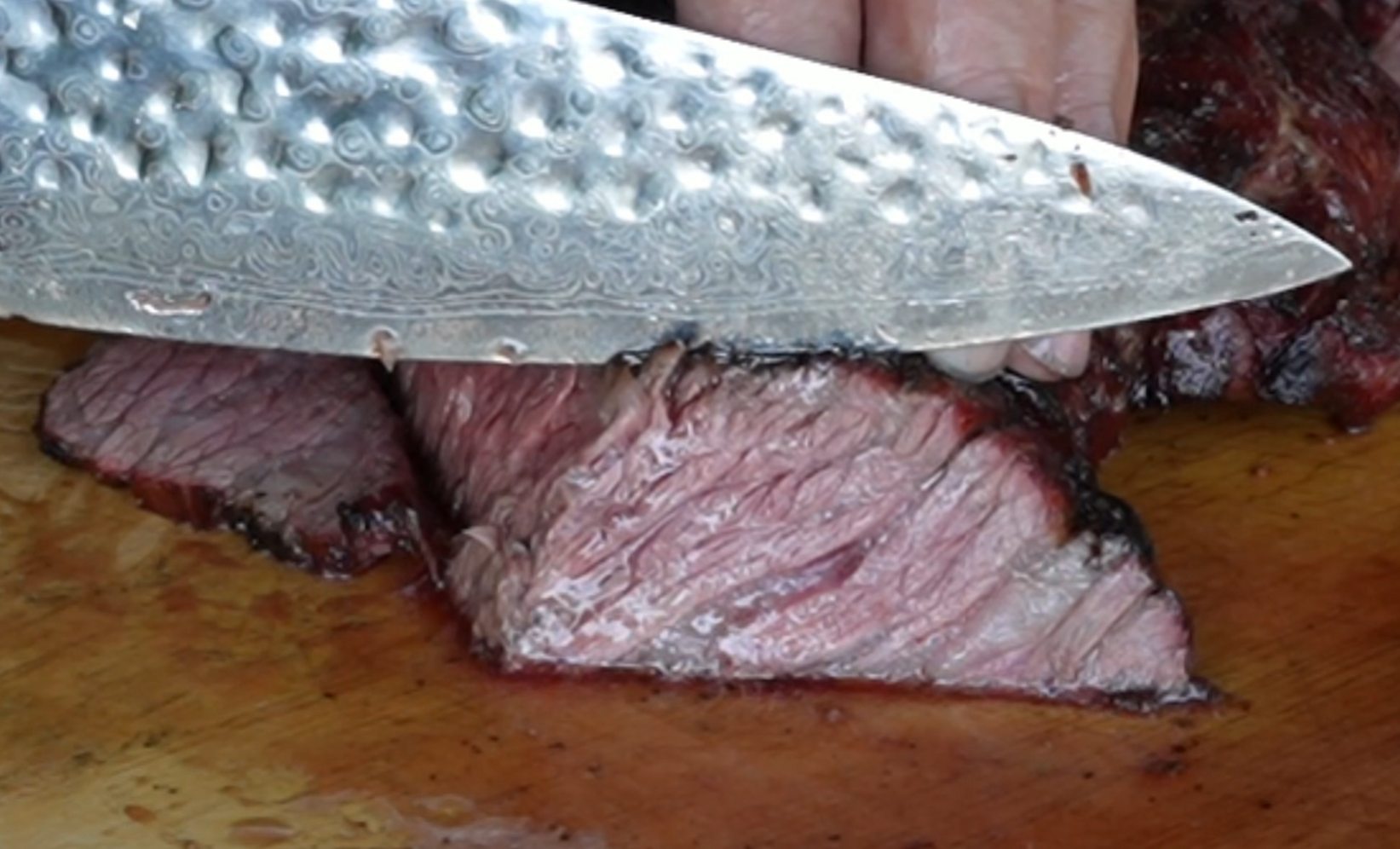 A close-up of a large knife slicing through a juicy, cooked piece of beef on a wooden cutting board.