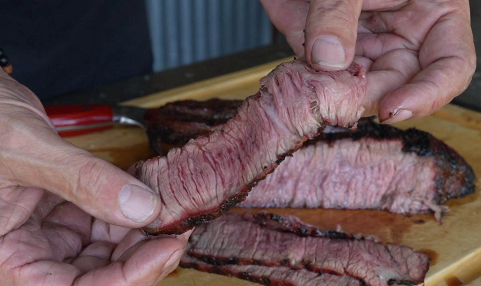 Hands holding a sliced piece of grilled steak showing a juicy, pink interior and charred exterior, with more sliced steak on a wooden cutting board in the background.