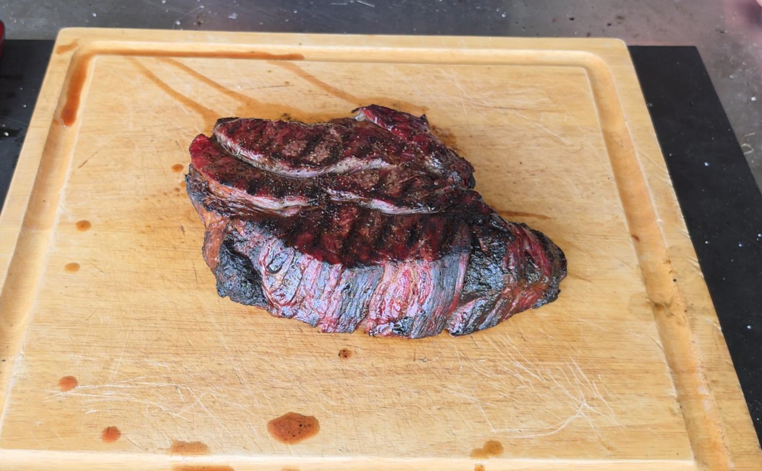 A piece of grilled steak with char marks rests on a wooden cutting board, surrounded by a few drops of meat juice.