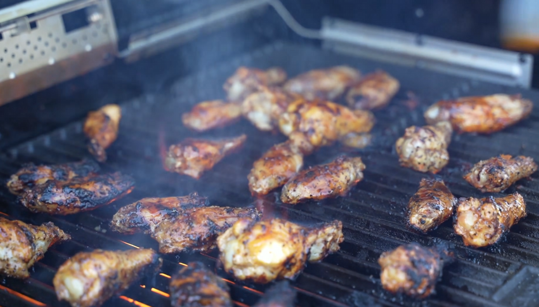 Chicken wings cooking on a grill, with smoke rising and grill marks visible on the wings. The grill appears to be outdoors, and the chicken is seasoned and slightly charred.