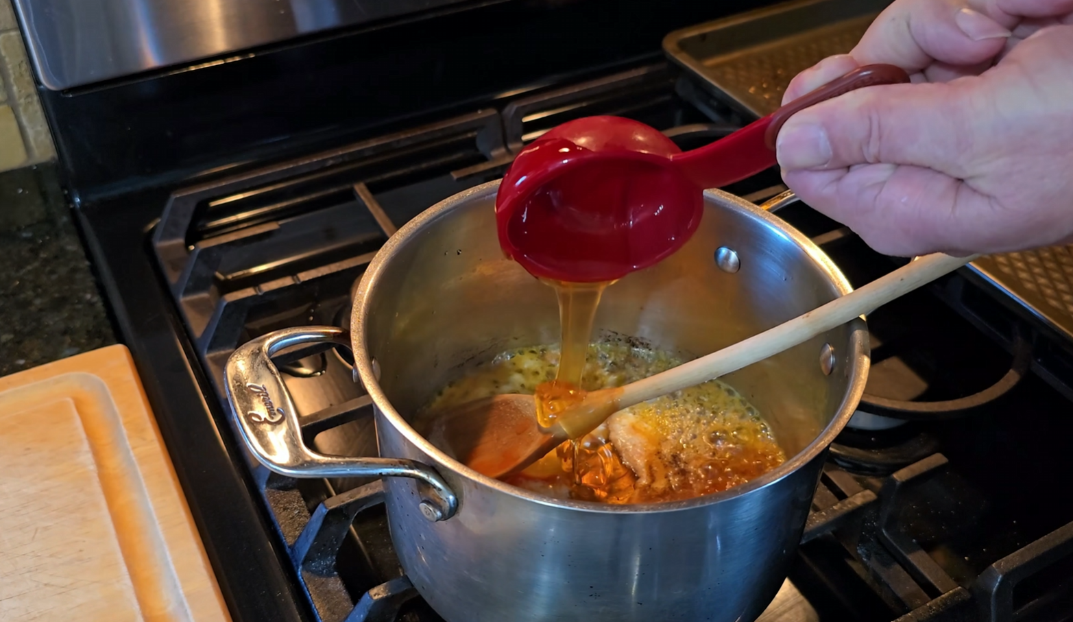 A person pours a brown liquid from a red measuring cup into a pot on a stove, stirring the mixture with a wooden spoon.