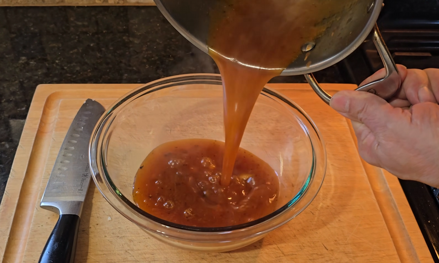 A person pours brown gravy with chunks from a pot into a clear glass bowl on a wooden cutting board. A large kitchen knife is placed next to the bowl.