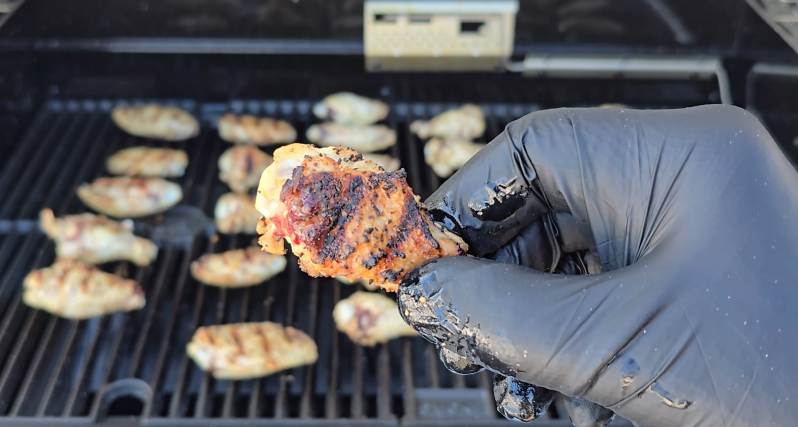 A gloved hand holds a grilled chicken wing in front of a barbecue grill filled with more wings.