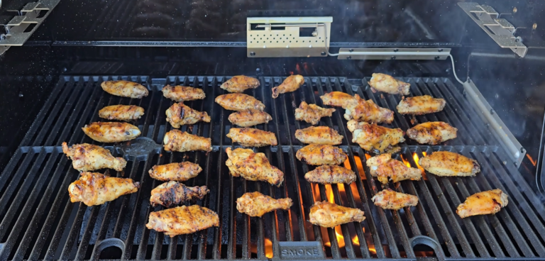 Chicken wings are grilling on a barbecue outdoors. The wings are spread across the grill grates, showing char marks and flames below. Smoke is rising, and a smoker box is positioned at the back of the grill.
