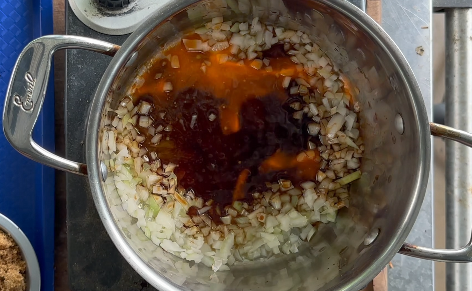 Chopped onions and a dark liquid sauce simmer in a stainless steel pot on a stovetop, with part of a blue cutting board and a container of brown sugar visible nearby.