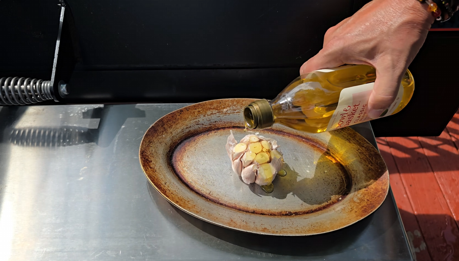A hand pours olive oil from a bottle onto a halved garlic bulb placed on a rustic oval metal plate, set on a shiny outdoor grill surface.