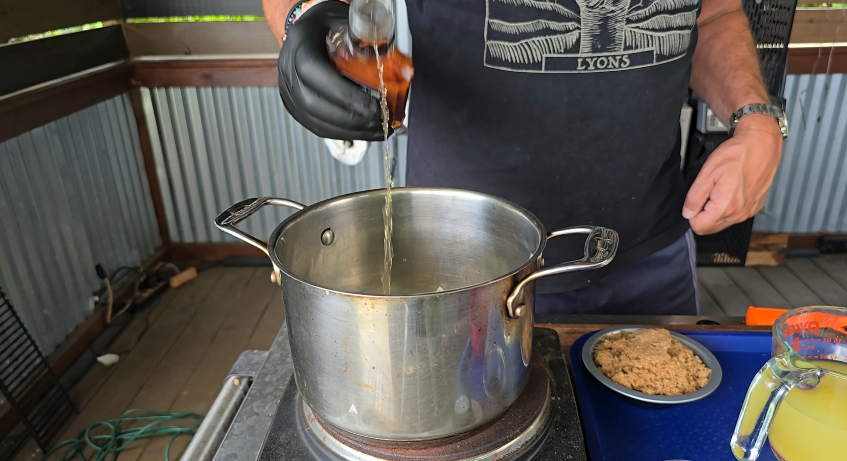 A person wearing a black glove pours liquid from a brown bottle into a large pot on a stove. Brown sugar and a measuring cup with yellow liquid sit nearby on a blue tray.