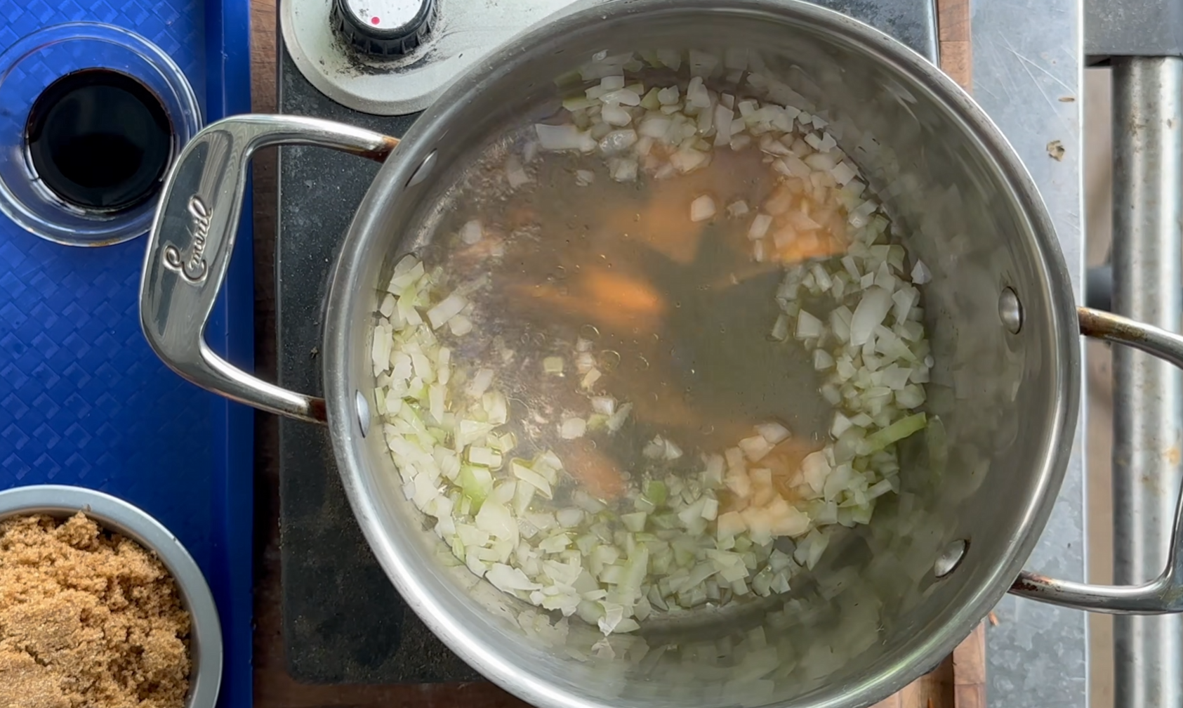 A pot on a stove contains chopped onions and carrots simmering in water. A small bowl of brown sugar and a cup of dark liquid are nearby on the counter.