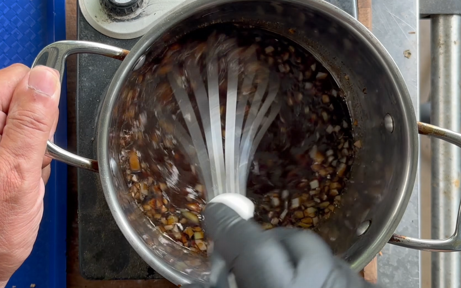 A person whisks a mixture of chopped onions and dark liquid in a metal pot on a stovetop, preparing a sauce or marinade.